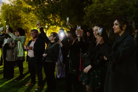 Grupo de personas con copas permanece de pie en un jardín al anochecer.