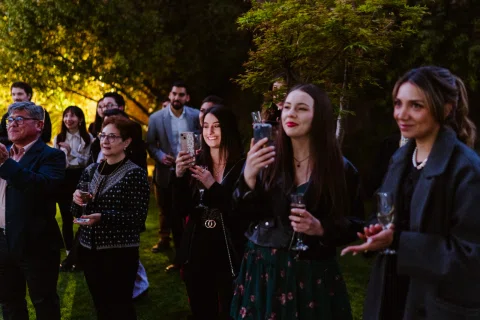 Grupo de personas permanece de pie con copas en un jardín al atardecer.