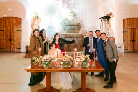 Grupo de personas posa alrededor de una mujer con vestido blanco sentada detrás de una mesa decorada.
