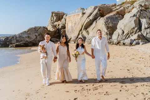 Grupo de personas posando junto en la arena con rocas y mar al fondo.
