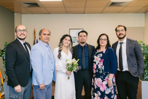 Grupo de personas posa en una sala alrededor de una mujer con vestido blanco y ramo.