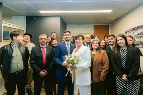 Grupo numeroso de personas posa dentro de una sala alrededor de una mujer con traje blanco y ramo.