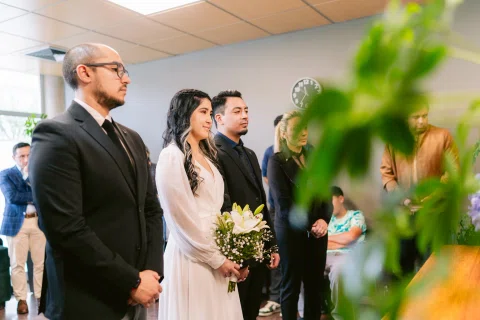Grupo de personas permanece de pie en una sala; una mujer con vestido blanco sostiene un ramo junto a un hombre vestido de negro.