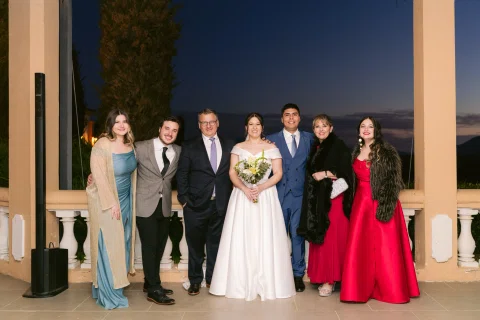 Grupo de siete personas posa al aire libre junto a una mujer con vestido blanco y ramo.