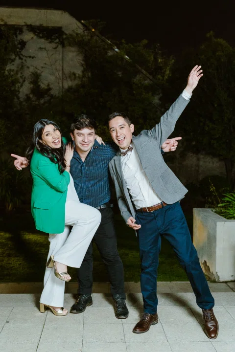 Tres personas posando con gestos de celebración en el jardín durante la noche.