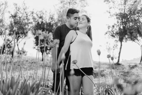 Hombre abraza desde atrás a una mujer entre plantas, en una fotografía en blanco y negro.