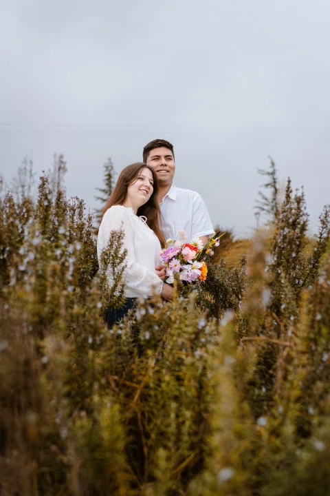 Hombre abraza a una mujer que sostiene un ramo entre vegetación alta.