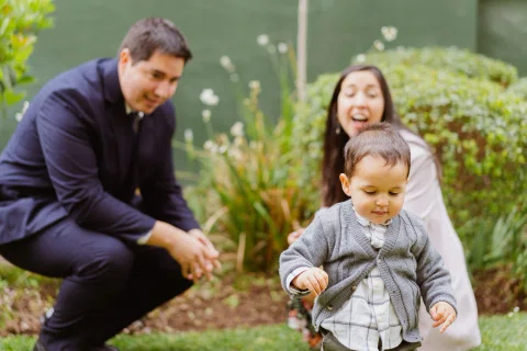 Hombre agachado extiende una mano hacia un niño pequeño mientras una mujer ríe detrás en un jardín.
