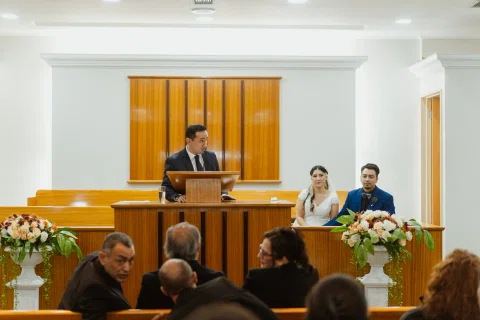 Hombre habla desde un atril mientras una mujer con vestido blanco y un hombre con traje azul permanecen sentados al fondo.