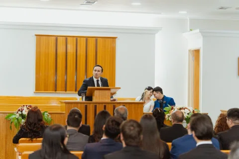 Hombre permanece detrás de un atril mientras varias personas están sentadas frente a él en una sala.