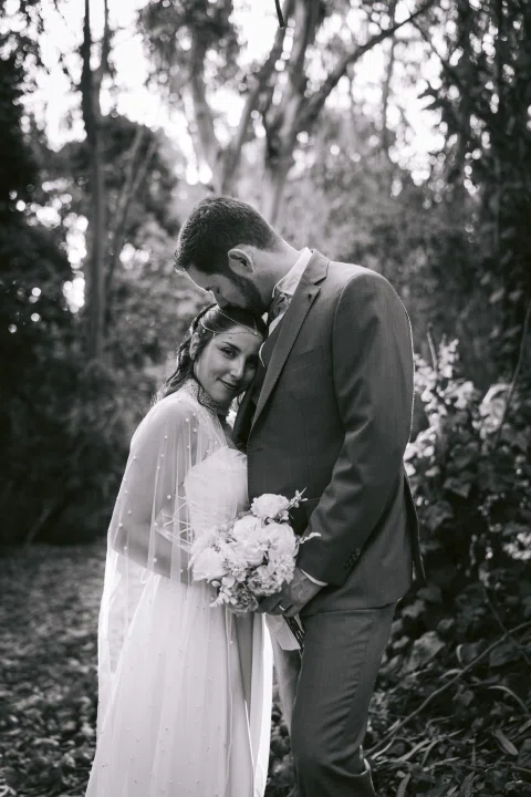 Hombre con traje oscuro besa la cabeza de una mujer con vestido blanco que sostiene un ramo, en una fotografía en blanco y negro.