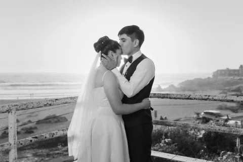 Hombre besa la frente de una mujer mientras ella sostiene su rostro, con la costa al fondo en blanco y negro.