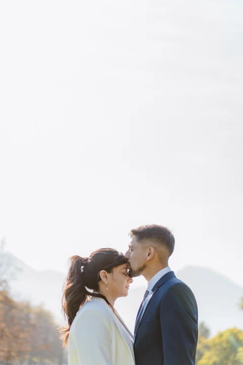 Hombre con traje azul besa la frente de una mujer con traje claro al aire libre, con montañas desenfocadas al fondo.
