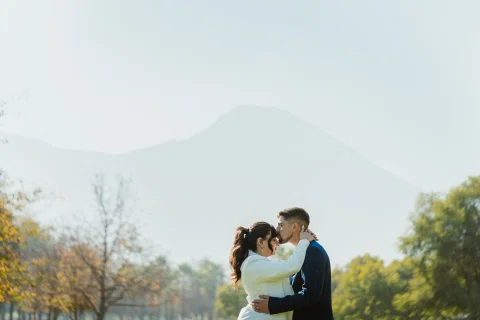 Hombre con traje azul besa la frente de una mujer con traje claro mientras la abraza, con una montaña al fondo.