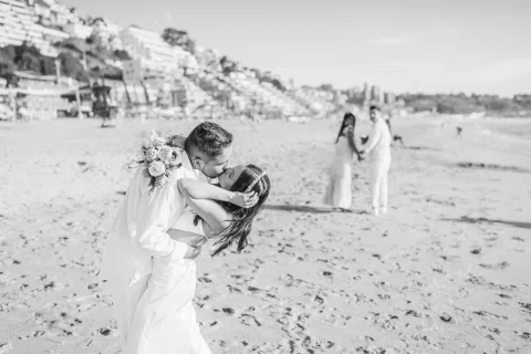 Hombre besa a una mujer inclinada hacia atrás en la playa, con otras dos personas desenfocadas al fondo, en blanco y negro.