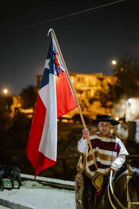 Hombre con poncho y sombrero monta a caballo mientras sostiene una bandera de Chile durante la noche.