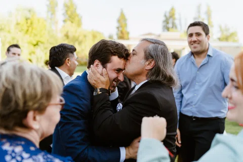 Hombre de cabello gris besa la mejilla de un hombre con traje azul al aire libre.
