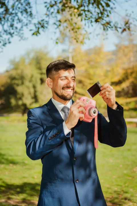 Hombre con traje azul sonríe mientras sostiene una cámara instantánea rosada y muestra una fotografía.