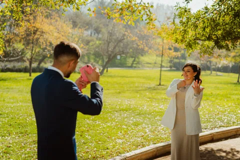 Hombre con traje azul fotografía con una cámara instantánea rosada a una mujer con traje claro que posa a distancia.