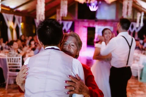 Hombre con camisa blanca abraza a una mujer vestida de rojo en la pista, con otras personas bailando al fondo.