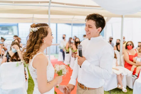 Hombre con camisa blanca sostiene un micrófono frente a una mujer con vestido blanco y ramo de flores.