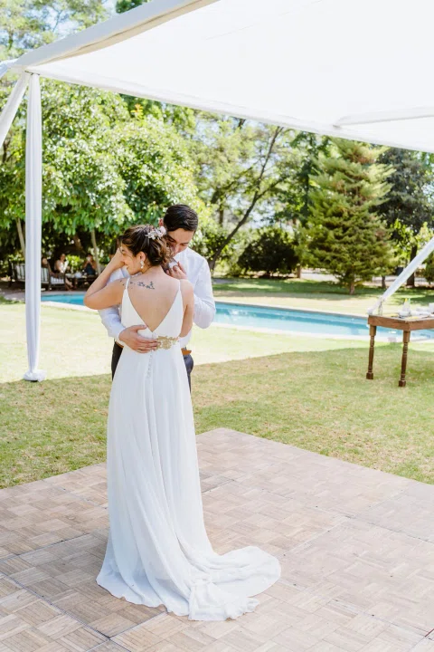 Hombre con camisa blanca y mujer con vestido blanco se abrazan bajo una carpa exterior.