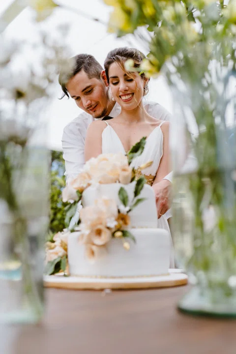 Hombre con camisa blanca abraza a una mujer con vestido blanco detrás de una torta decorada con flores.