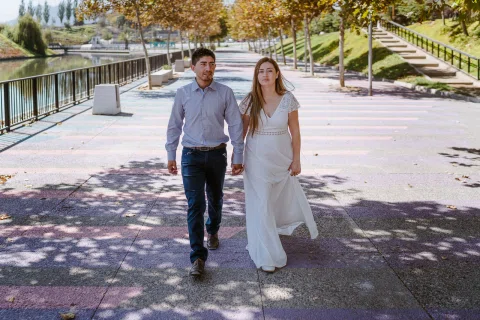 Hombre con camisa celeste y mujer con vestido blanco caminan juntos por un sendero de un parque.