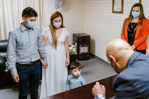 Hombre con camisa celeste y mujer con vestido blanco están junto a un niño frente a un hombre con traje y mascarilla.