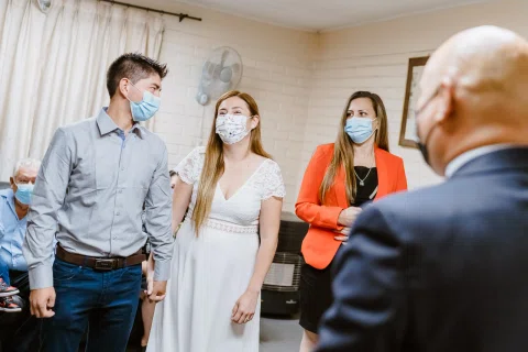 Hombre con camisa celeste y mujer con vestido blanco están frente a un hombre con traje y mascarilla dentro de una sala.