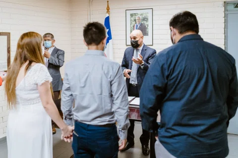 Hombre con camisa celeste y mujer con vestido blanco están frente a un hombre con traje negro dentro de una sala.