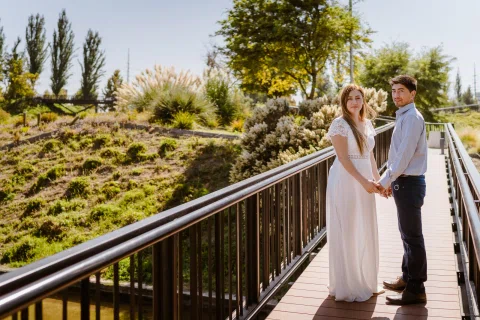 Hombre con camisa celeste y mujer con vestido blanco posan juntos junto a la baranda de un puente.