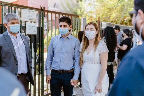 Hombre con camisa celeste y mujer con vestido blanco, ambos con mascarilla, de pie en el exterior junto a otras personas.