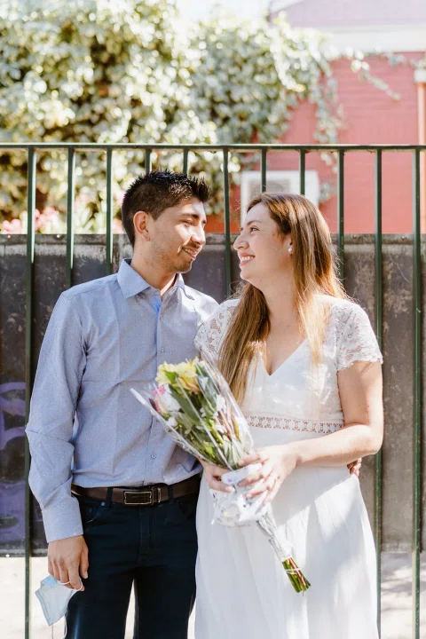 Hombre con camisa celeste y mujer con vestido blanco y ramo posan juntos junto a una reja.