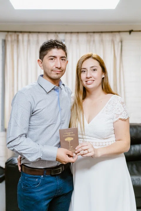 Hombre con camisa celeste y mujer con vestido blanco posan juntos sosteniendo una libreta marrón.