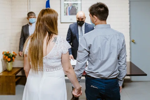 Hombre con camisa celeste y mujer con vestido blanco vistos de espaldas y tomados de la mano frente a un hombre con traje.