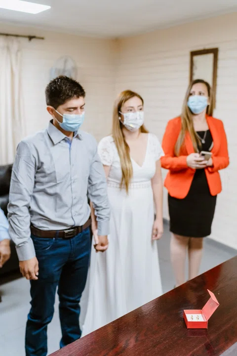 Hombre con camisa celeste y mujer con vestido blanco están de pie y tomados de la mano junto a una mesa; una mujer con blazer rojo permanece detrás.
