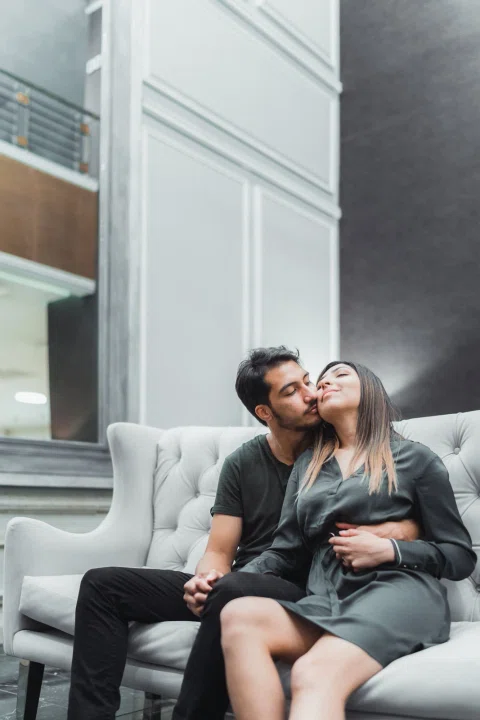 Hombre con camiseta oscura besa la frente de una mujer con vestido oscuro mientras ambos están sentados en un sofá.