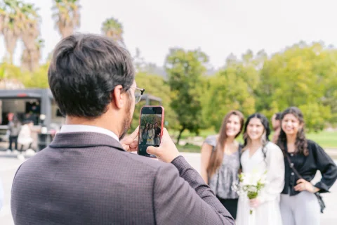 Hombre de espaldas sostiene un celular frente a tres mujeres que posan al aire libre.