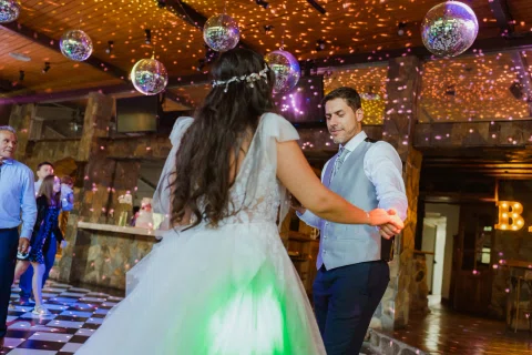 Hombre con chaleco y mujer con vestido blanco bailan tomados de las manos bajo bolas de espejo y luces de colores.