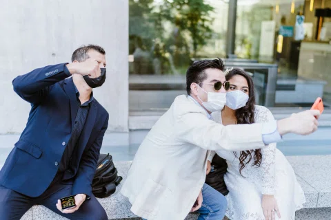 Hombre con chaqueta blanca toma una selfie junto a una mujer con vestido blanco y mascarilla, mientras otro hombre aparece a un lado.