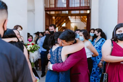 Hombre con chaqueta burdeos abraza a una mujer con vestido azul entre varias personas en el exterior de la iglesia.