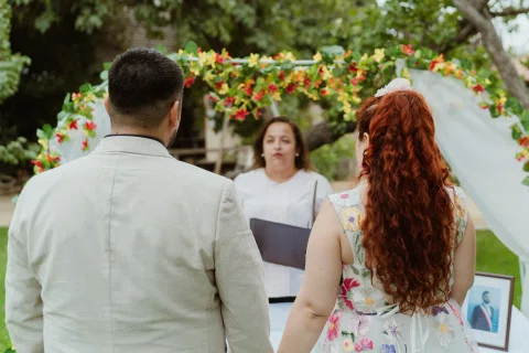 Hombre con chaqueta clara y mujer de cabello rojo permanecen de espaldas frente a una mujer con vestido blanco bajo un arco floral.