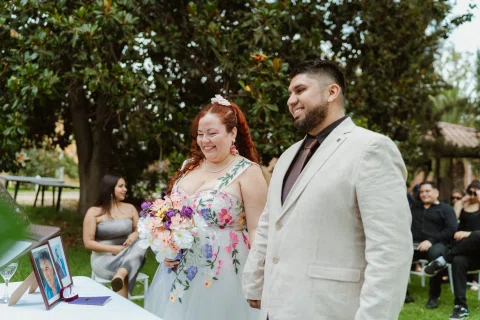 Hombre con chaqueta clara y mujer de cabello rojo posan juntos; ella sostiene un ramo colorido.