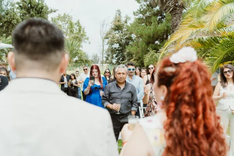 Hombre con chaqueta clara y mujer de cabello rojo miran a un hombre que se acerca, con varias personas al fondo.
