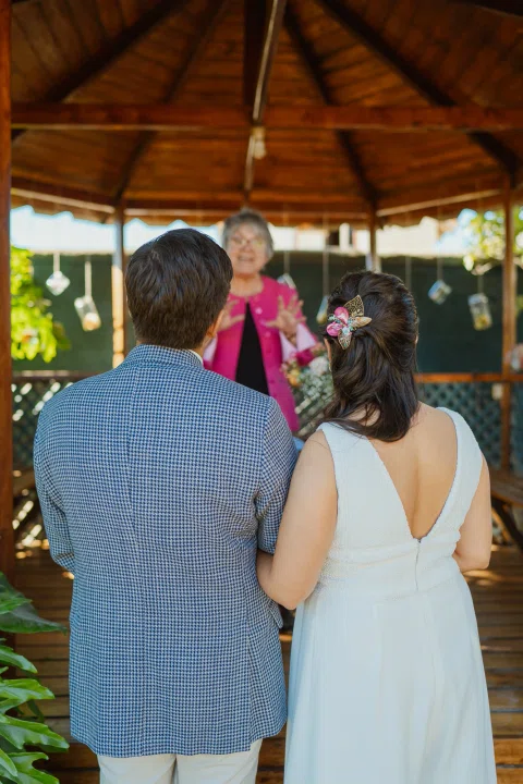 Hombre con chaqueta clara y mujer con vestido blanco permanecen frente a una mujer con carpeta dentro del gazebo.
