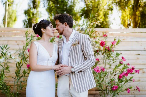Hombre con chaqueta a rayas y mujer con vestido blanco se abrazan junto a arbustos con flores rosadas.