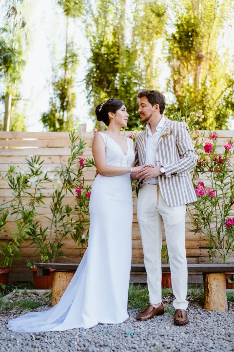 Hombre con chaqueta a rayas y mujer con vestido blanco posan de cuerpo entero frente a un muro de madera y flores.