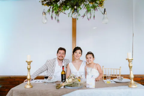 Hombre con chaqueta a rayas, mujer con vestido blanco y una niña están sentados juntos en una mesa con velas y bebidas.