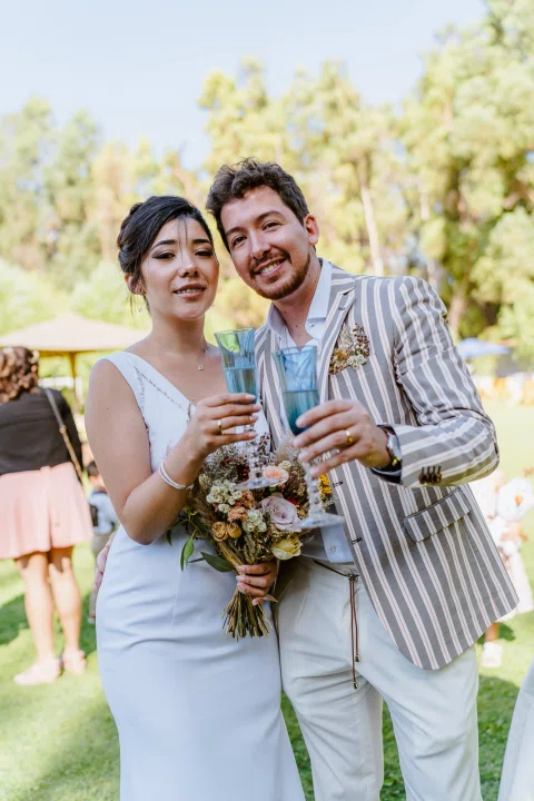 Hombre con chaqueta a rayas y mujer con vestido blanco posan juntos; ella sostiene un ramo de flores.